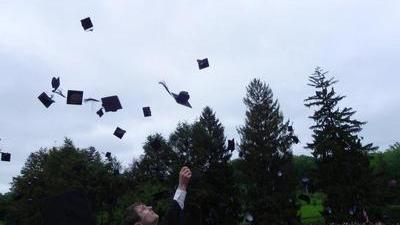 graduates at an outside commencement throwing their hats in the air, with pine trees in the background