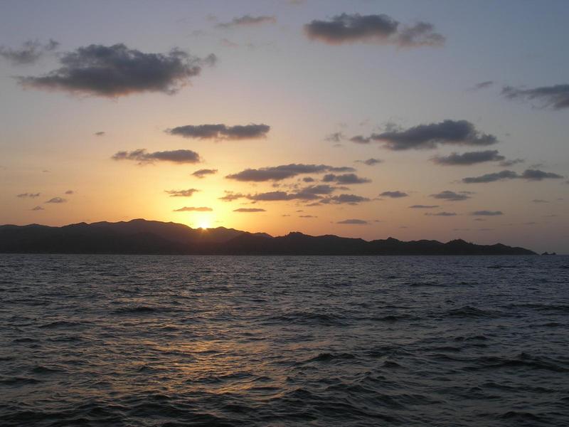 a glow of yellow sun shrinks behind rocky hills across dark ocean water, puffs of clouds lit from the back in the sky