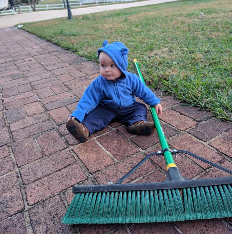toddler sitting on a brick path by a lawn looking sideways, holding a large green pushbroom