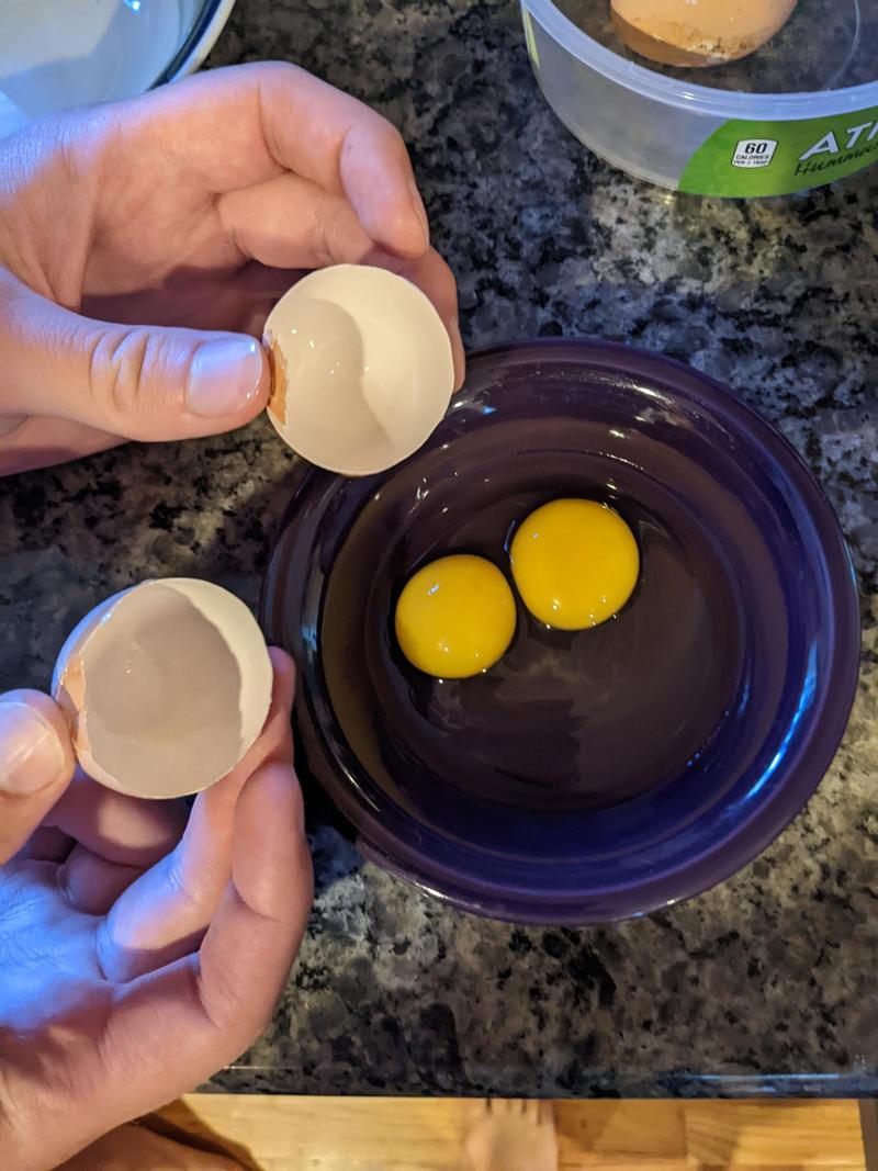 hands holding open two halves of an egg shell, a ceramic bowl underneath cradling the white and two small yolks