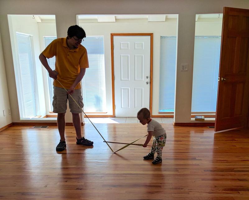 Randy and his toddler son measuring an empty room with a tape measure as sunlight streams in the window onto the hardwood floors