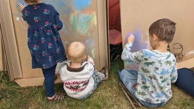 Three young children with their backs to the camera coloring with chalk pastels on large pieces of cardboard