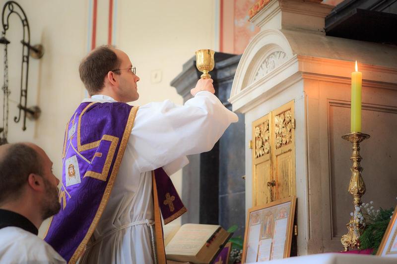 A Catholic priest in purple and gold holds up a chalice in front of an altar