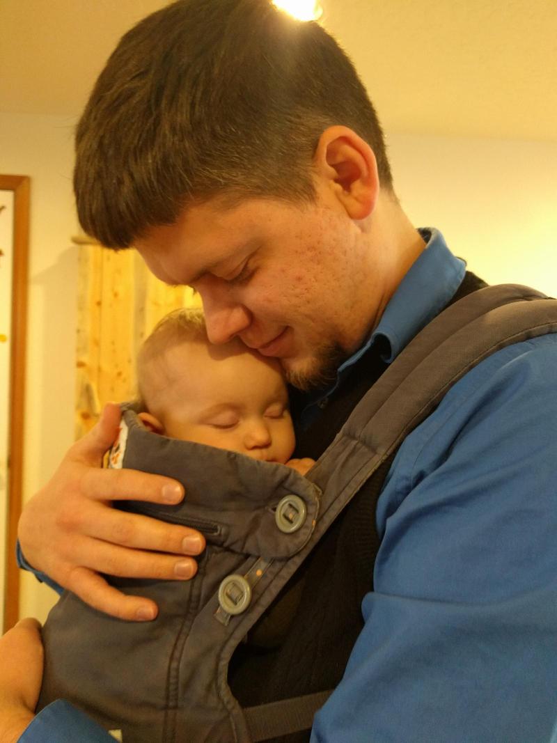 Randy wearing and hugging baby in an ergo baby carrier, leaning his head down to touch the baby's head and smiling