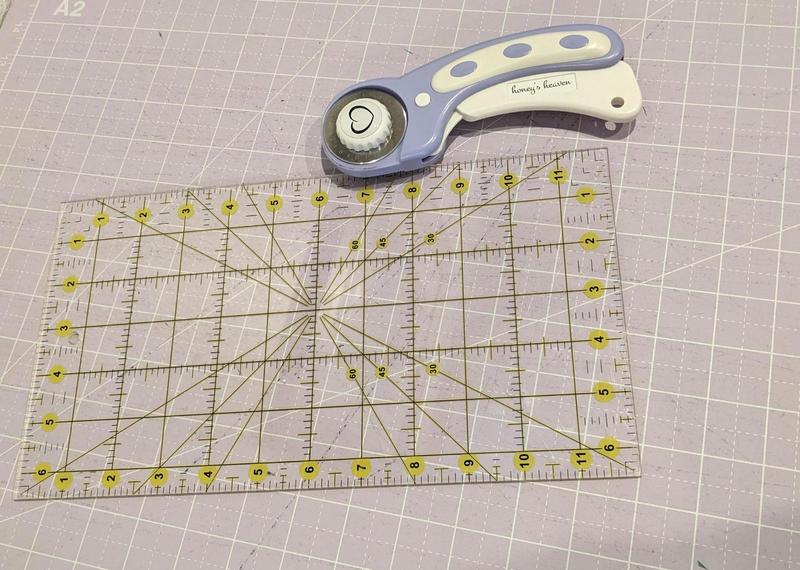 a rotary cutter resting on a cutting mat on a table beside a plastic rectangle marked with yellow lines for measuring cuts