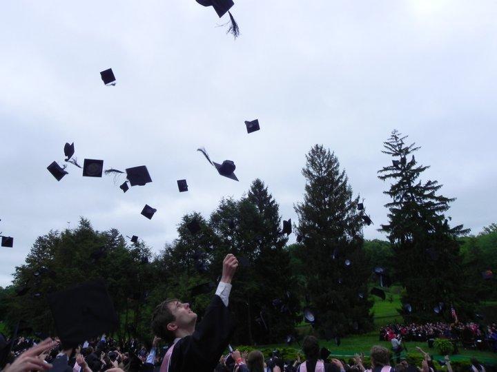 graduates at an outside commencement throwing their hats in the air, with pine trees in the background